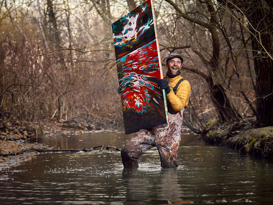 Smiling artist holds abstract painting while wading in creek.